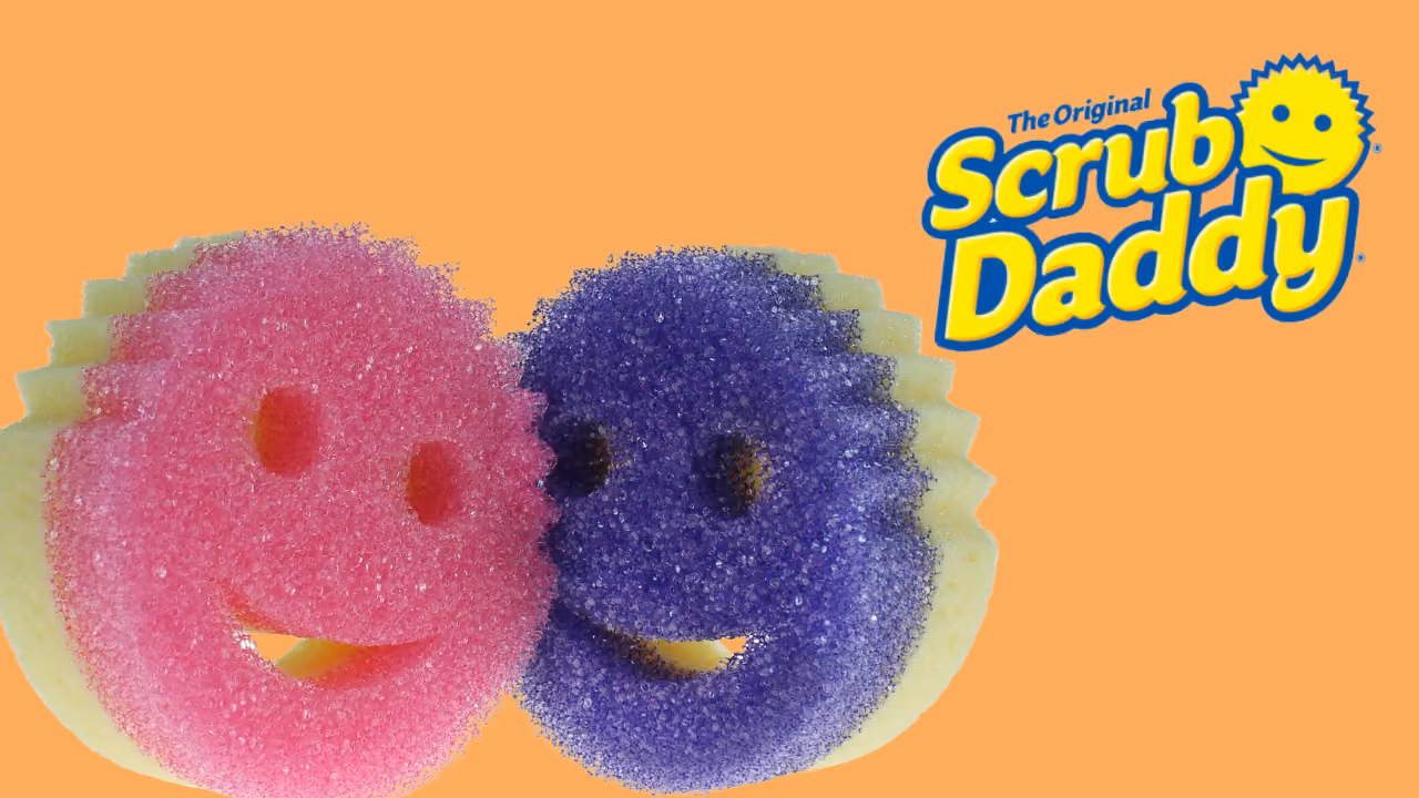 Two Scrub Daddy sponges, pink and purple, overlapping beside the logo on orange