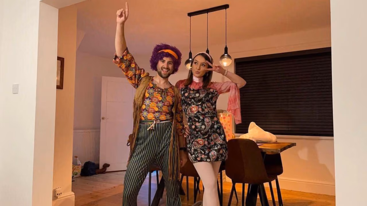 Two people in 1960s fancy dress in a dining room — a purple afro wig, floral shirt and striped flares; a paisley mini dress and a peace sign