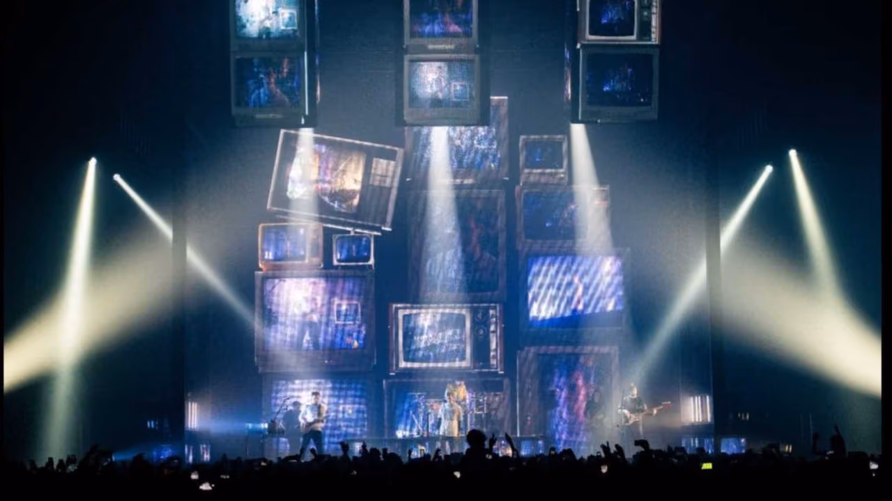 A band on stage under a tower of stacked old televisions, white spotlights raking across a crowd of raised phones