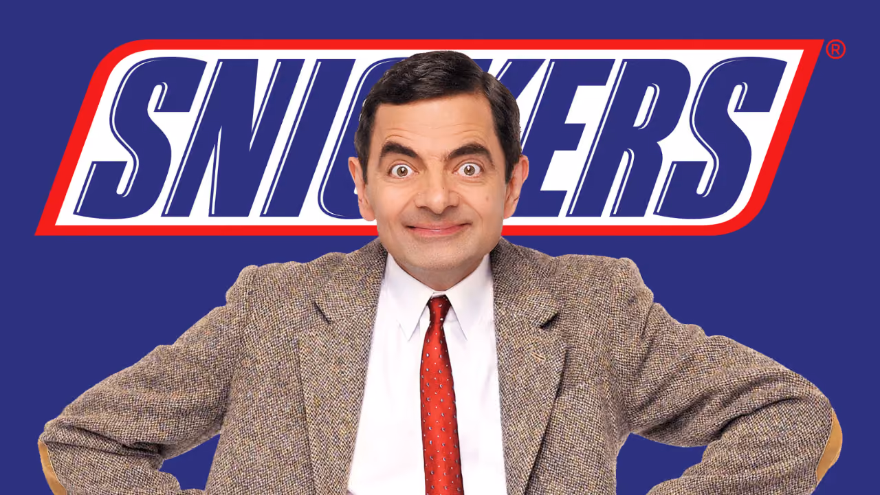 Rowan Atkinson as Mr Bean, hands on hips, in front of the Snickers logo on deep blue