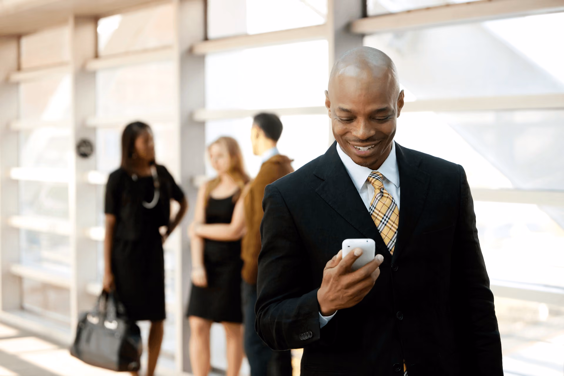 A man in a dark suit and gold tie smiling at his phone in a bright glass-walled foyer, three colleagues talking behind him