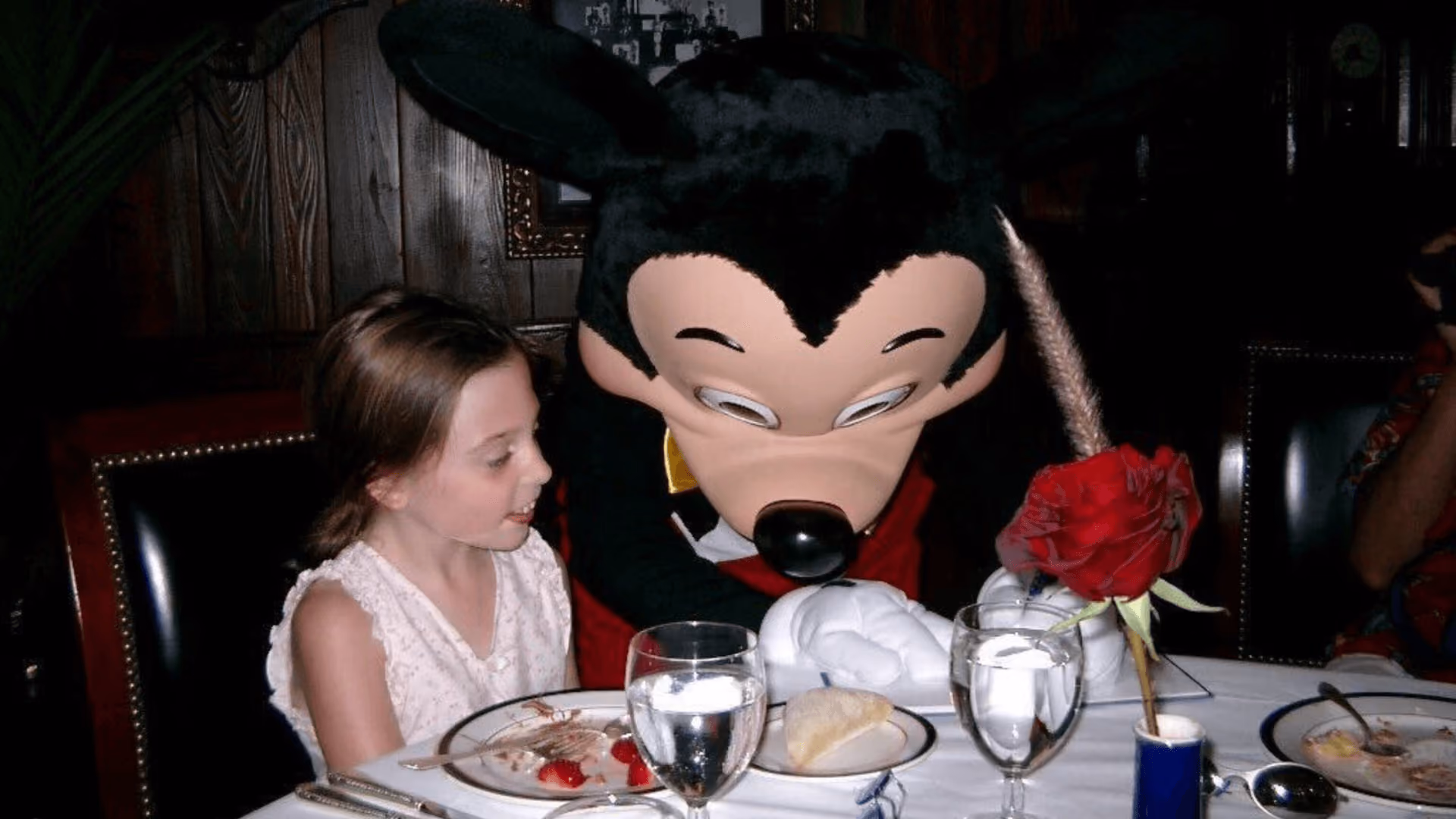 A girl at a restaurant table looking up at Mickey Mouse in black tie, who is leaning in beside her, a red rose in a vase on the tablecloth