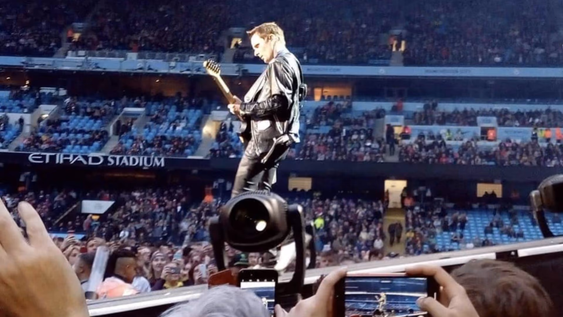 A guitarist in a leather jacket playing on a walkway out into the crowd at the Etihad Stadium, the stands full behind him and phones raised below