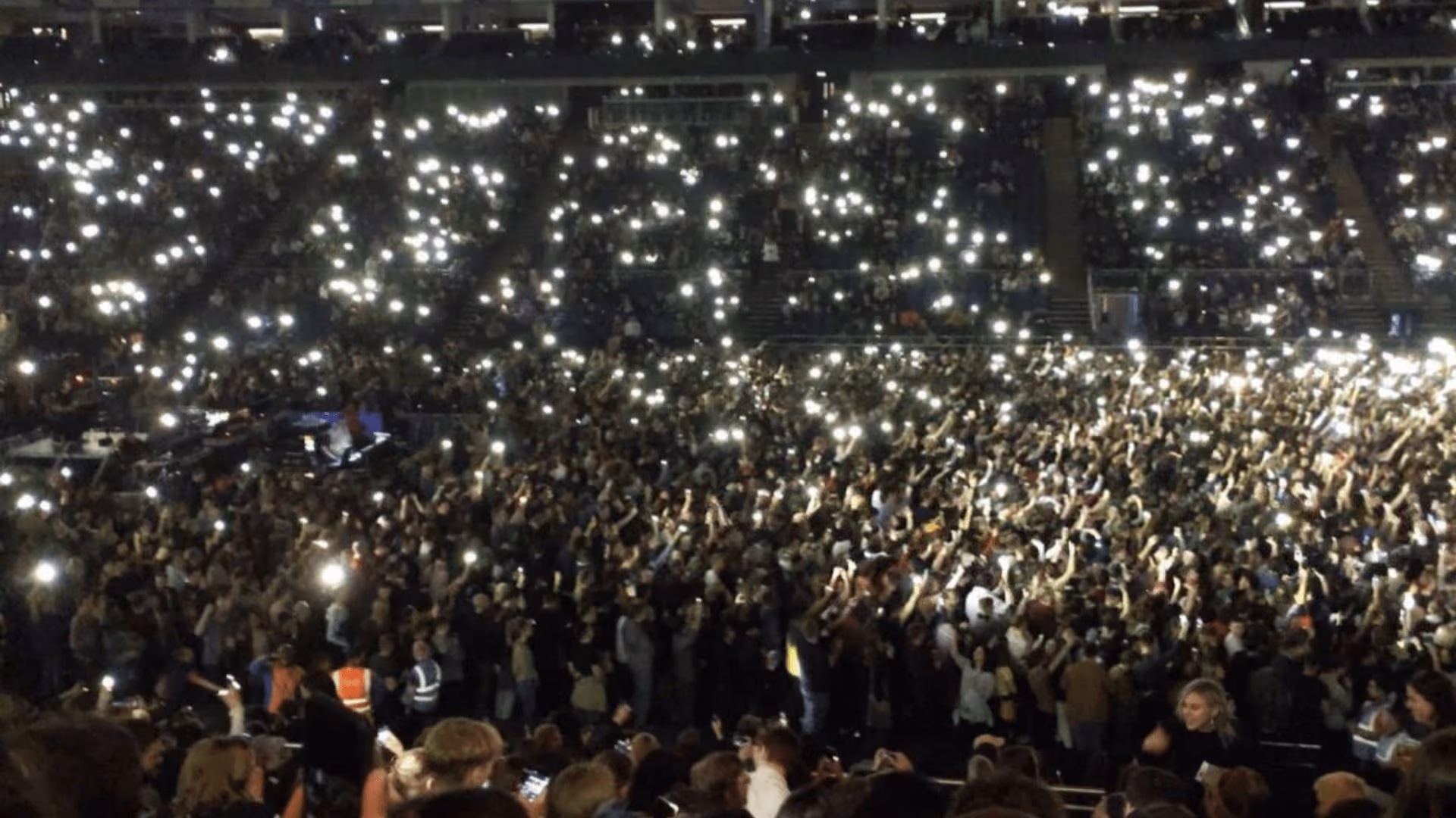 A stadium crowd at night seen from high up, thousands of phone torches held aloft across the floor and the tiers