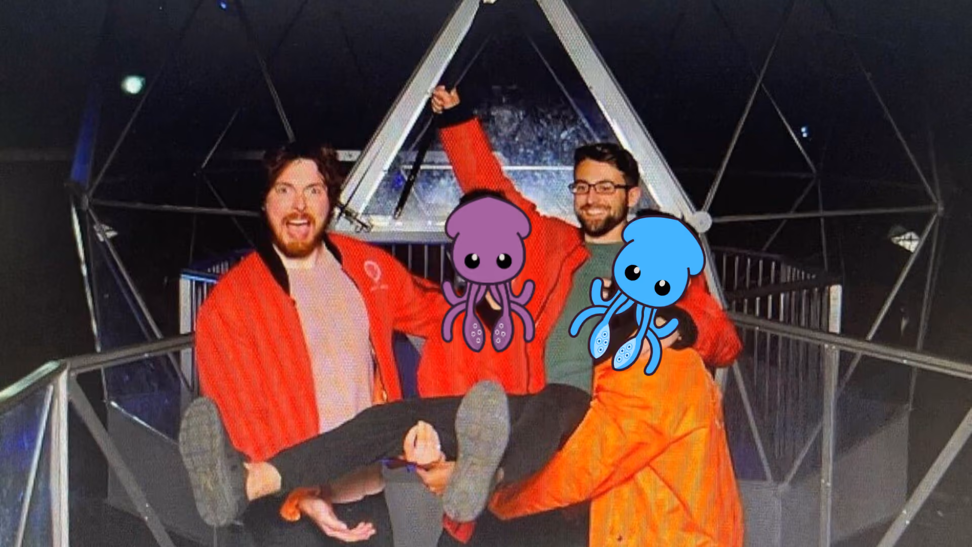 Three people in Crystal Maze jumpsuits inside the geodesic crystal dome, two of them carrying a third, two faces covered by purple and blue cartoon squid