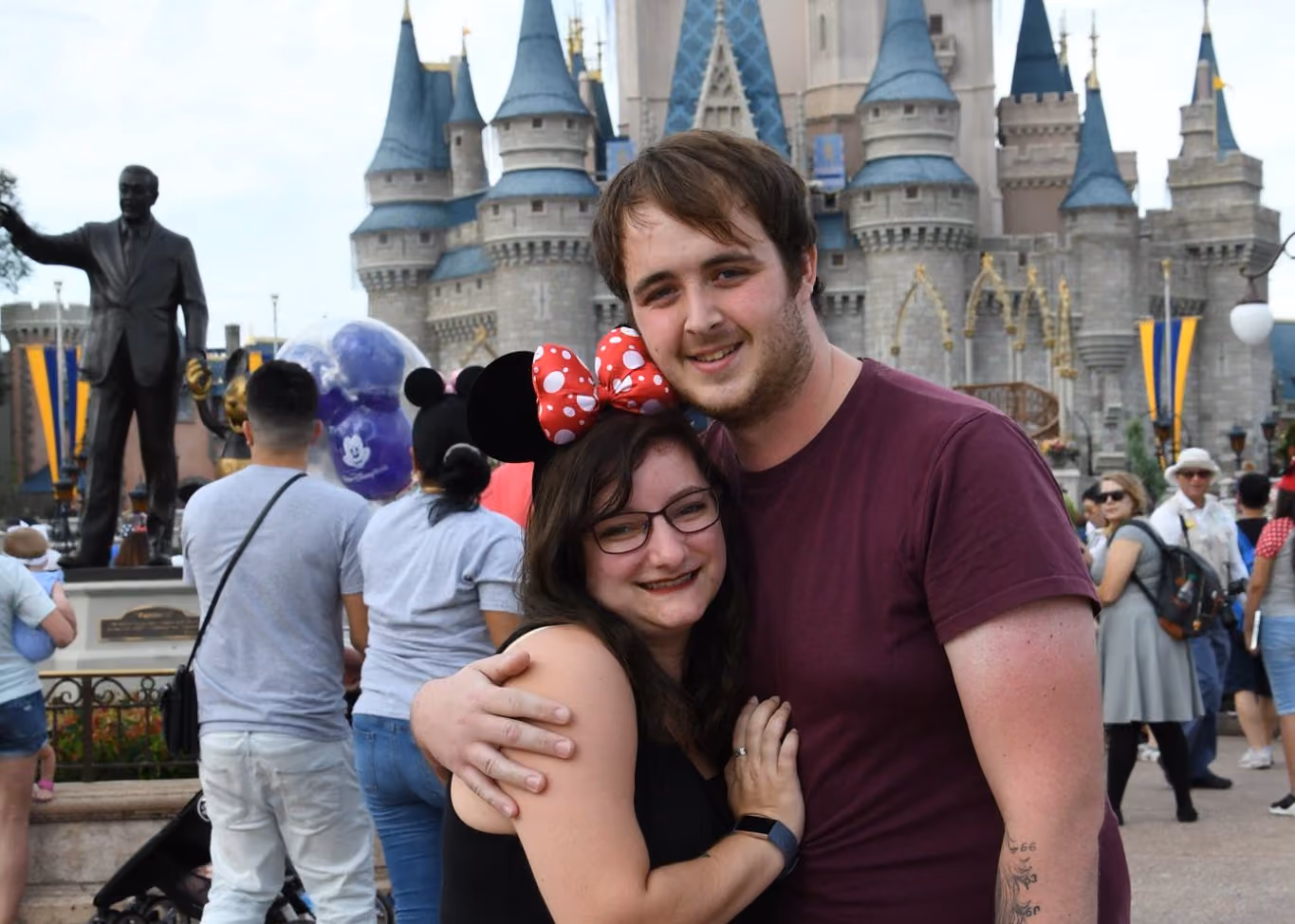 A couple in front of Cinderella Castle at Walt Disney World, she in Minnie Mouse ears with a red spotted bow, the Partners statue of Walt Disney and Mickey behind them