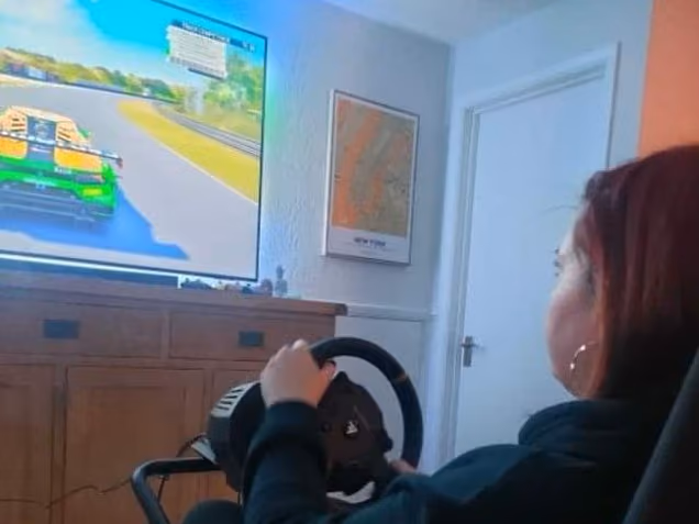 Seen over her shoulder, a woman at a racing wheel playing a driving game on a wall-mounted television