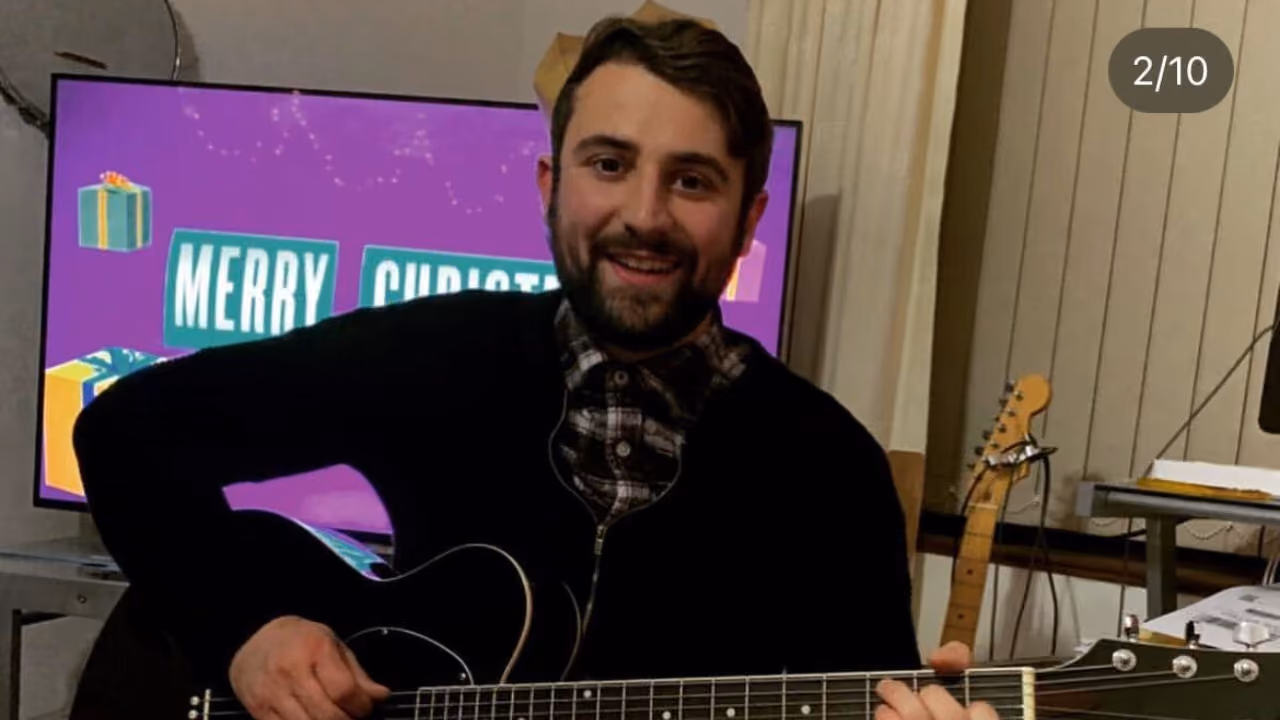 A man in a paper crown playing an electric guitar in front of a television showing a Merry Christmas graphic, a second guitar on a stand behind him