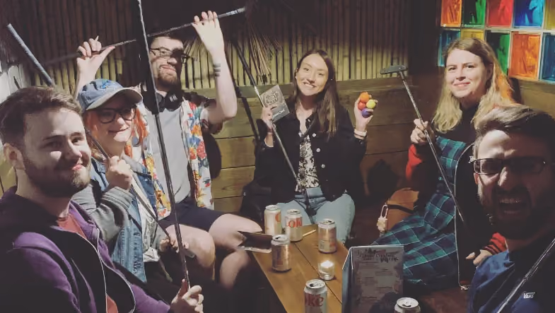 Six colleagues round a table in a tiki-themed bar, each holding a putter aloft, drinks and a scorecard in front of them