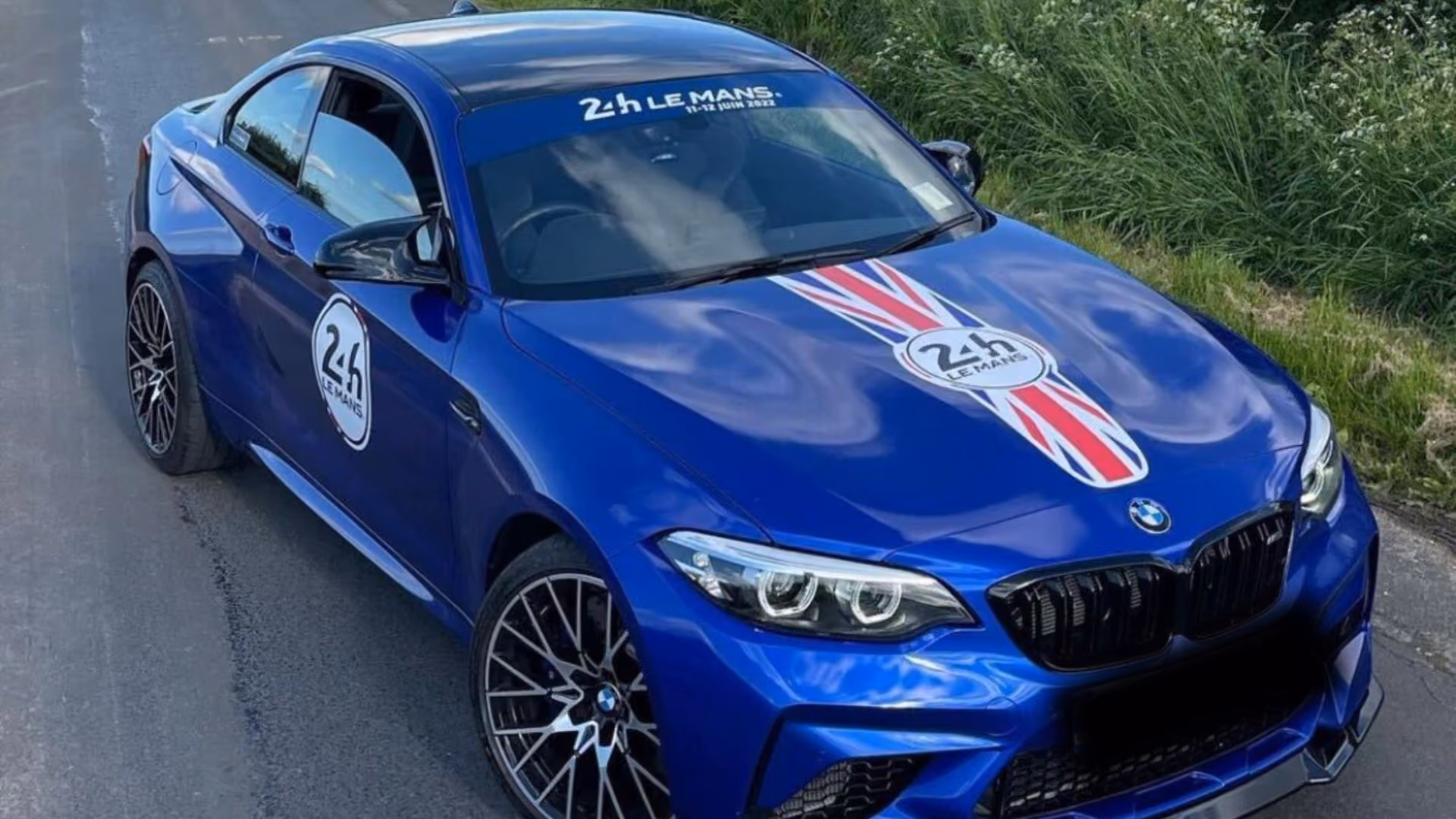 A blue BMW M2 on a country lane, decalled with 24h Le Mans roundels and a Union flag stripe down the bonnet