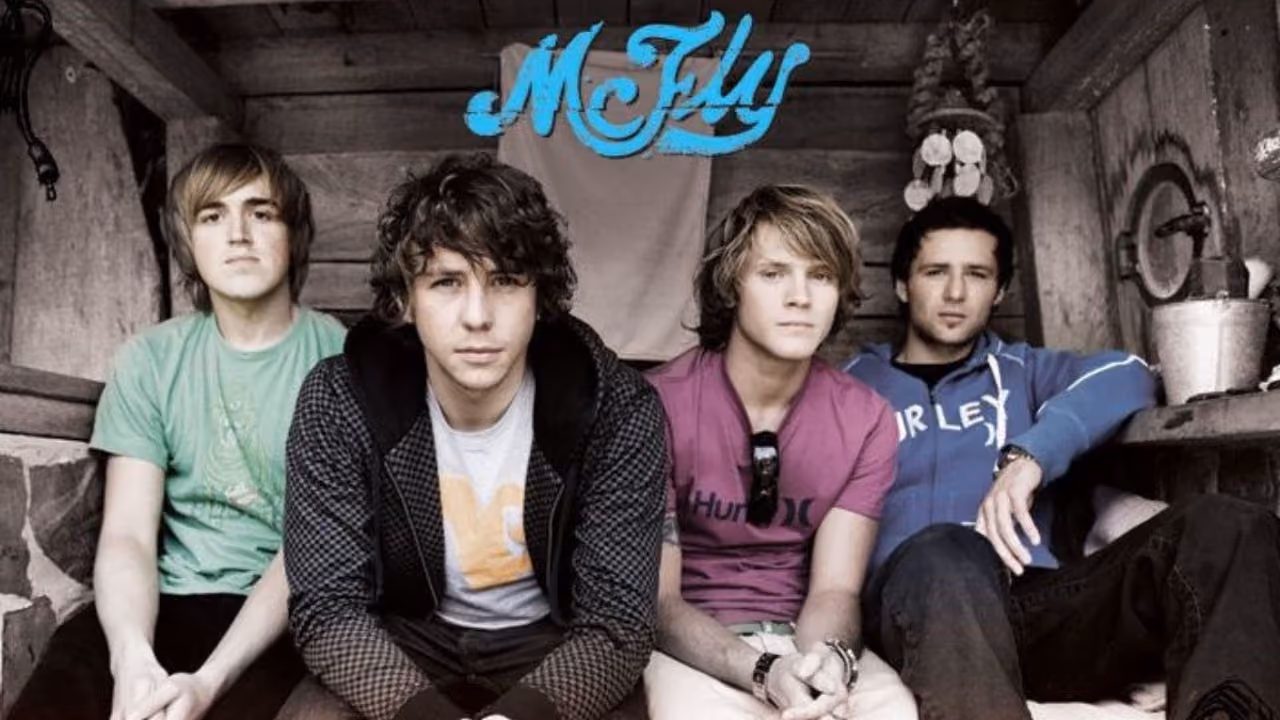 A McFly promotional photograph, the four of them sitting against the wooden wall of a cabin, the band's logo above them in blue