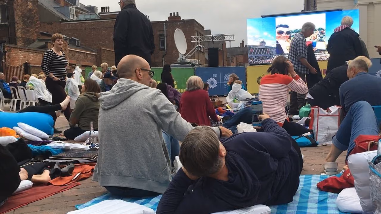 An open-air screening in a car park: people on blankets, cushions and airbeds on the tarmac watching a big screen, brick buildings and a satellite truck behind