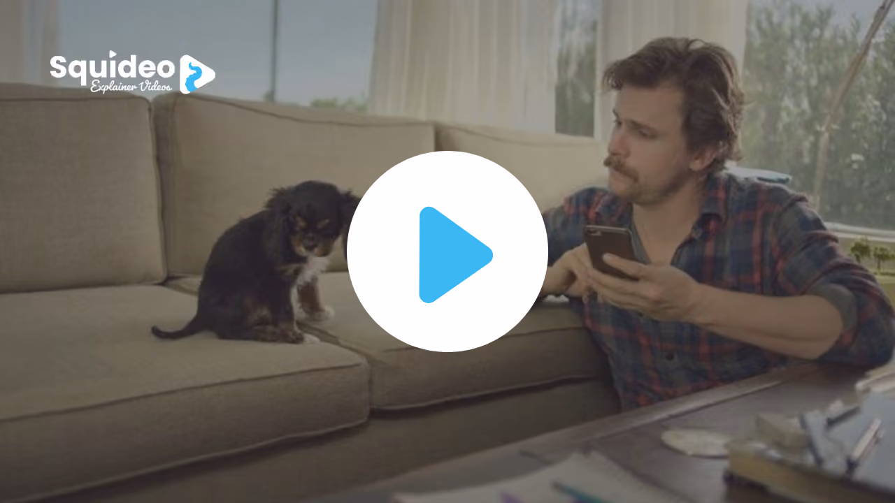 A man in a checked shirt sitting on the floor with his phone, a small black and tan puppy on the sofa beside him