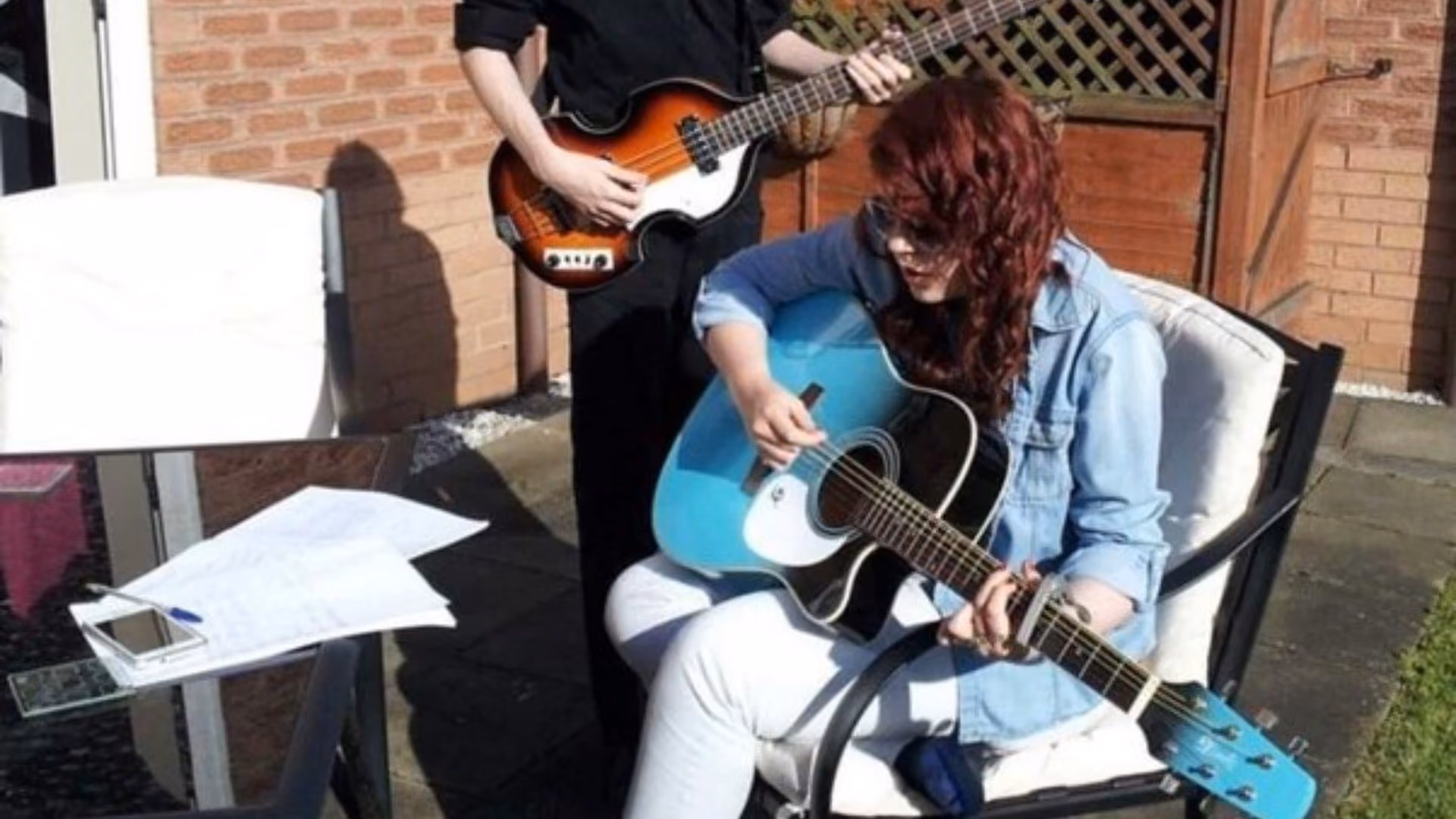 Two people playing in a back garden — one standing with a sunburst bass, the other sitting in a denim jacket with a pale blue acoustic guitar, handwritten sheets on the table beside them