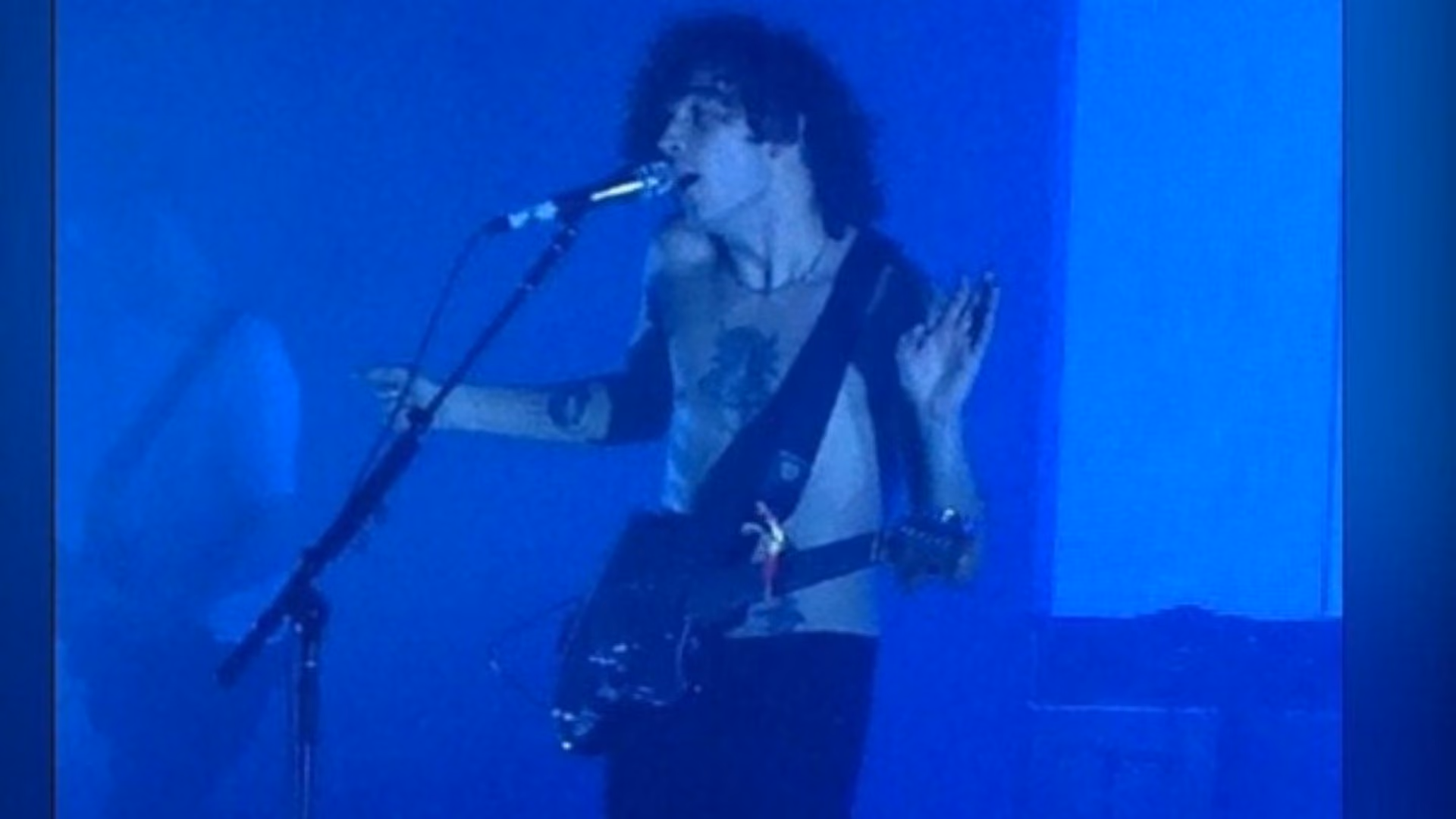 A guitarist singing into the microphone under deep blue stage light, one hand raised