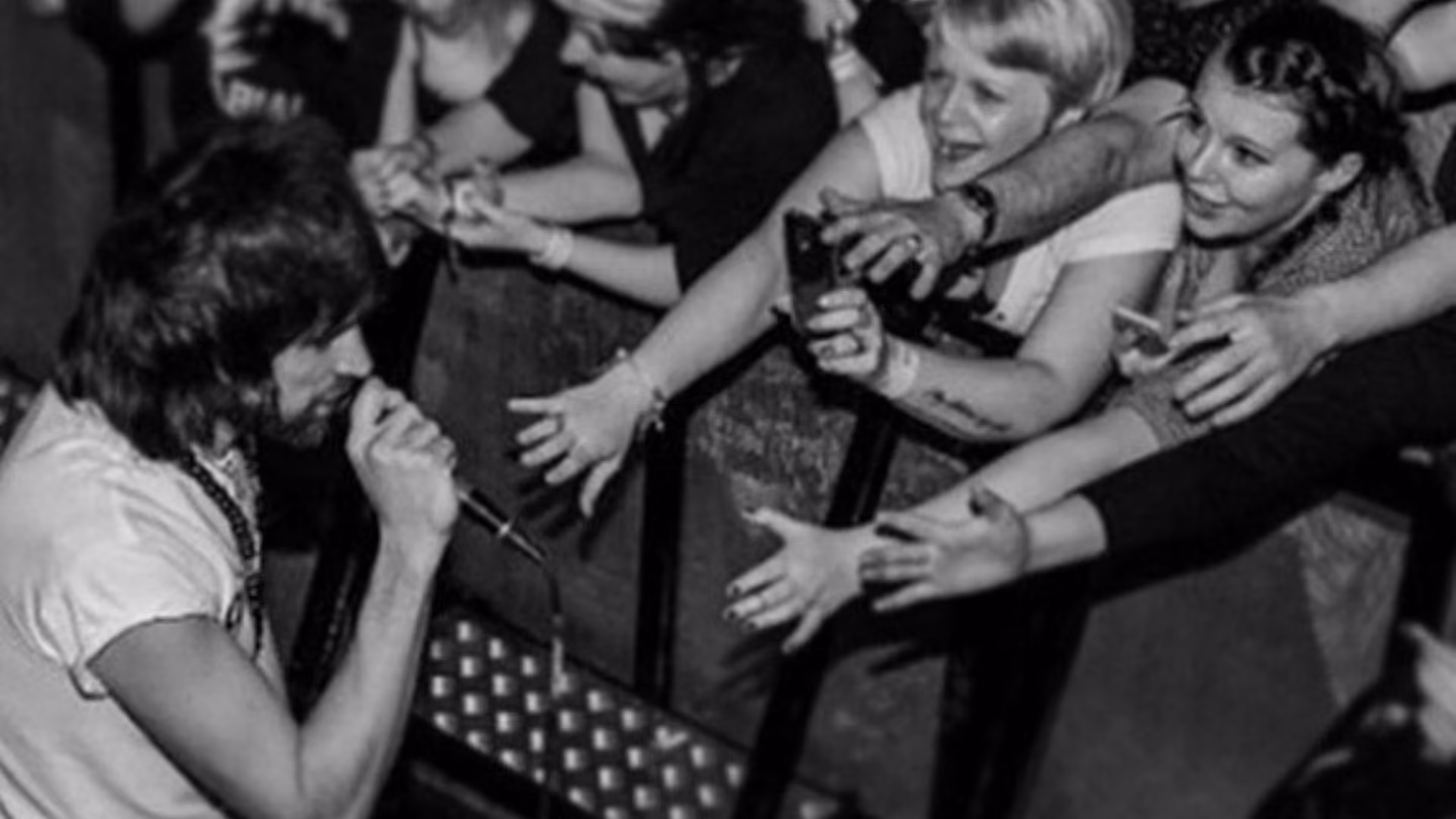 Black and white: a musician leaning down from the stage to sign for fans stretched over the barrier, phones held out towards him