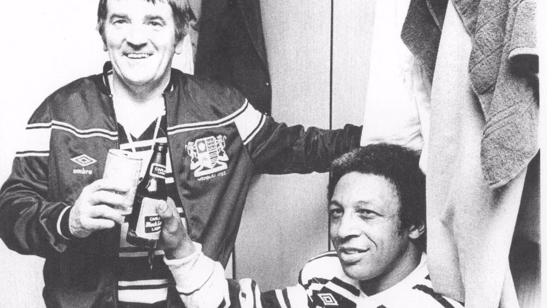 A black and white dressing-room photograph: a man in a club tracksuit top raising a glass, another player beside him holding a bottle of Carling Black Label, a Wembley 1982 crest on the jacket