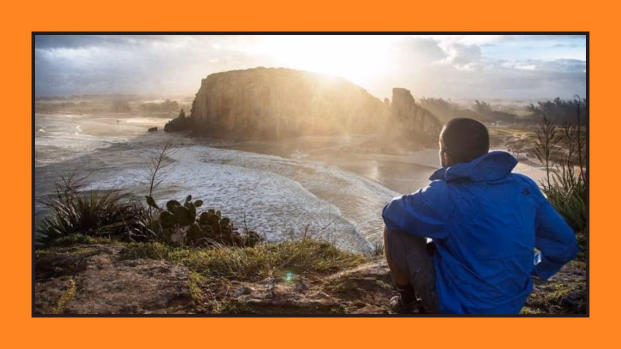 A man in a blue North Face jacket sitting on a headland at sunrise, looking out over a beach and a rocky outcrop