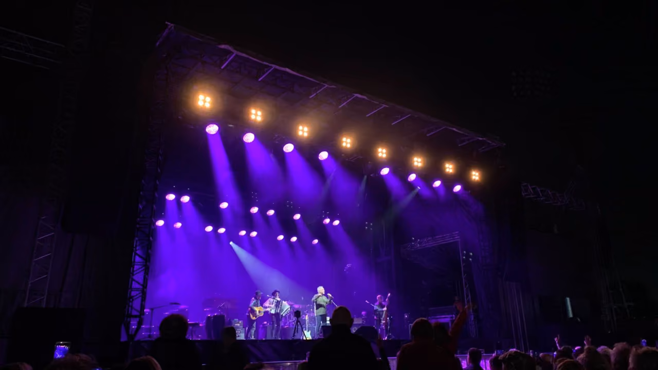An outdoor concert at night, the band small on a wide stage under banks of purple and white lights, heads of the audience in silhouette